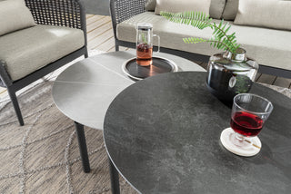 Das DeVries Lounge-Set Lanzarote Rope grau umfasst zwei runde Steintische mit einem Glas Rotwein, Krug und Vase, umgeben von geflochtenen Stühlen mit Rope-Geflecht. Das Set wird durch sein elegantes Aluminium-Gestell für eine perfekte Mischung aus Stil und Komfort aufgewertet.