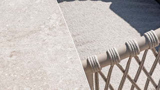 Eine Nahaufnahme einer beige strukturierten Oberfläche, die an das Latte-Keramik-Finish des 4Seasons-Tischs Prado erinnert, neben einem mit Seilen umwickelten Geländer, das zarte Schatten auf den sandigen Boden wirft.