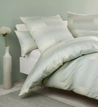 Ein minimalistisches Schlafzimmer mit einem weißen Bett in Janines Jersey-Bettwäsche Carmen S 55105-06 Salbeigrün, vor hellgrünen Wänden und einer Vase mit einer weißen Blume schafft eine Atmosphäre zeitlosen Designs.