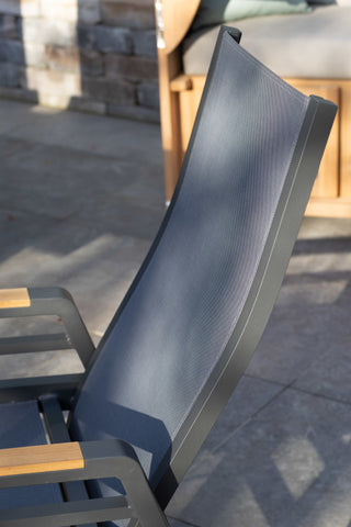 Der DeVries Dining-Sessel Montana Alu/Textilene verfügt über eine elegante dunkelgraue Netzrückenlehne und Holzarmlehnen sowie einen robusten Aluminium-Rahmen, der für Langlebigkeit auf der Steinfliesenoberfläche sorgt.