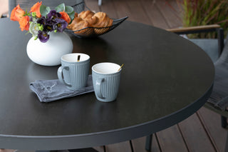 Zwei Becher mit Löffeln stehen auf dem DeVries Dining-Tisch Livingston Ø120cm, dazu eine Serviette und eine weiße Vase mit bunten Blumen. Croissants in einem Korb werten dieses charmante, moderne Design auf.
