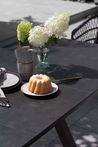 Ein Gugelhupf steht auf einem DeVries Ausziehtisch Hammond-Tisch, umgeben von Hortensien in einer Vase und einer gestreiften Tasse, was eine malerische Szene schafft.