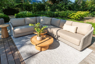 Auf einer Außenterrasse wird das Zeo Living 4Seasons Lounge-Set Casanova Rope Latte präsentiert, das aus einem L-förmigen beigen Sofa, runden Holztischen und einer Topfpflanze auf einem grauen Teppich besteht. Ergänzt wird es durch wetterfeste Kissen und üppiges Grün.