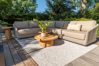 Auf einer Außenterrasse steht das 4Seasons Lounge-Set Casanova Rope Latte von Zeo Living, bestehend aus einem gewebten beigefarbenen Sofa mit wetterfesten Kissen, runden Holztischen und einem grauen Teppich. Dieses gemütliche Set ist wunderschön umgeben von leuchtend grünen Pflanzen, die zwischen den Bäumen eingebettet sind.