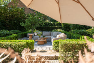 Das Zeo Living 4Seasons Lounge-Set Casanova Rope Latte umfasst ein beiges Sofa mit wetterfesten Kissen, Holztischen und einem Sonnenschirm, alles perfekt zum Entspannen inmitten üppigen Grüns.