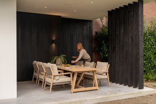 Eine Frau deckt elegant einen Tisch auf einer überdachten Terrasse mit den langlebigen Teakholzmöbeln von 4Seasons und farbenfrohen Topfpflanzen. Der hochwertige 4Seasons-Ess-Stuhl Julia aus Teakholz verleiht der gemütlichen Atmosphäre Eleganz.