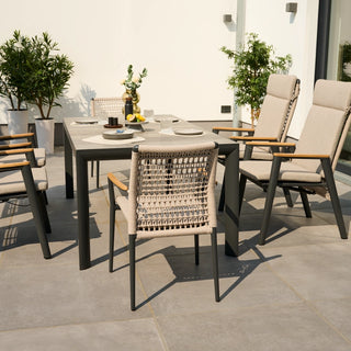 Garten-Essgruppe bestehend aus sechs Siena Garden Stapelstühlen Cortina natur mit Kissen, einem Tisch mit Geschirr und Topfpflanzen auf einer sonnigen Terrasse. Das Aluminium-Gestell wird elegant durch FSC-Teakholz-Elemente ergänzt.