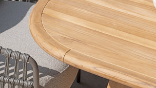 Nahaufnahme eines 4Seasons Tisch Capri 240 x 110 cm aus Teakholz mit abgerundeten Kanten neben einem grau gepolsterten Stuhl mit Metallrahmen, der Stil und Haltbarkeit zur Schau stellt.